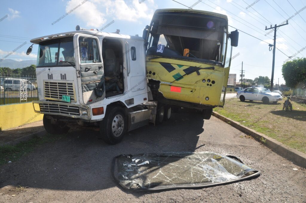 White cab-over truck with severe front-left damage after a collision with a yellow coach bus; shattered windshield on pavement; police vehicle in the background.