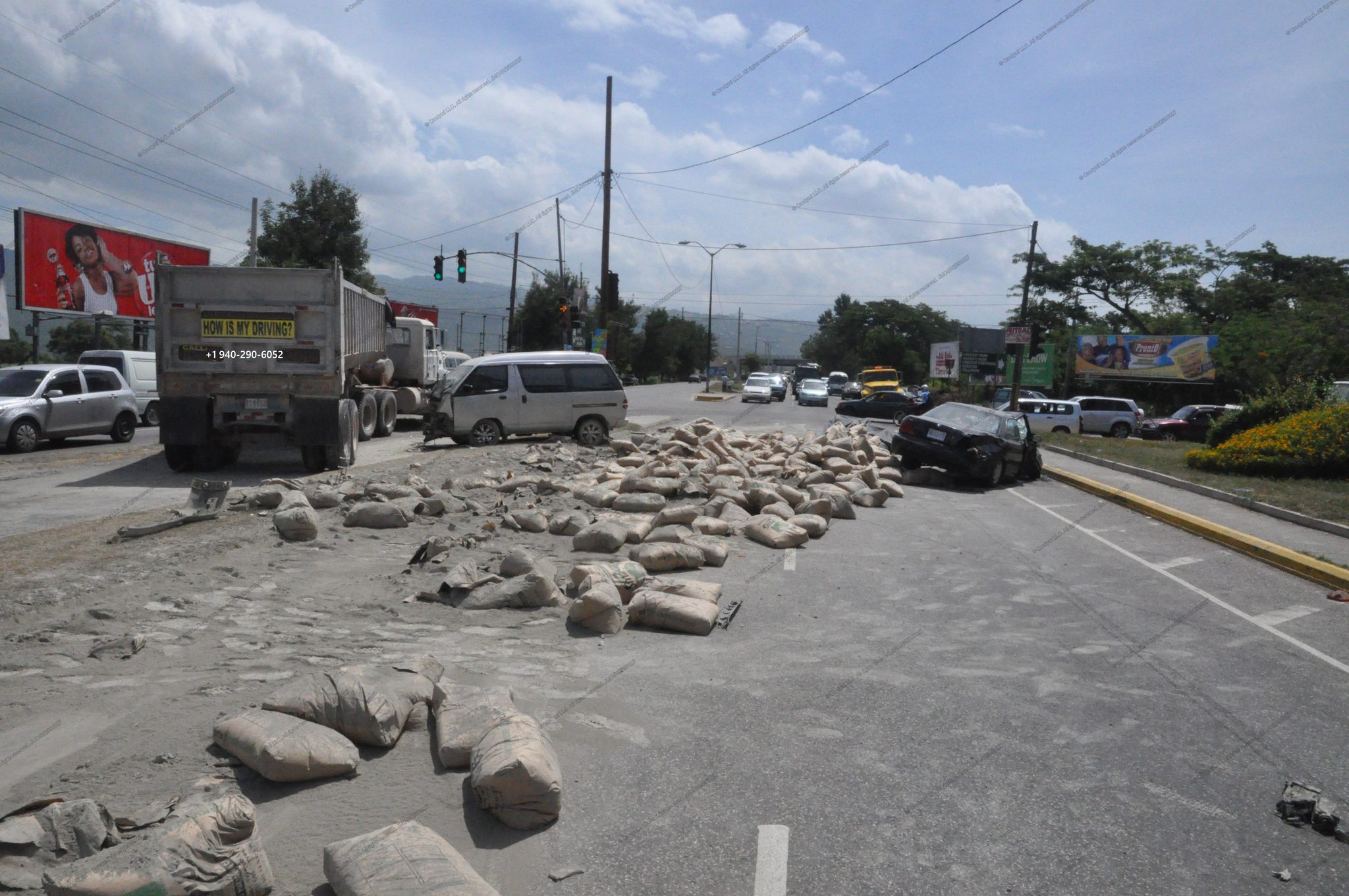 Cement bags spilled across multiple lanes after a dump-truck incident; white van stopped ahead and a damaged black sedan on the right shoulder at a busy intersection.